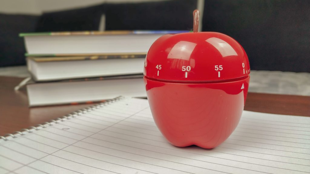 Closeup of pomodoro study technique kitchen timer in shape of a red apple for timing the studying with notebook and books in background laying on wooden desk