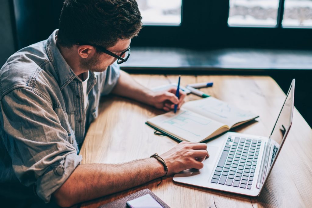 young man writing essay in notebook and laptop