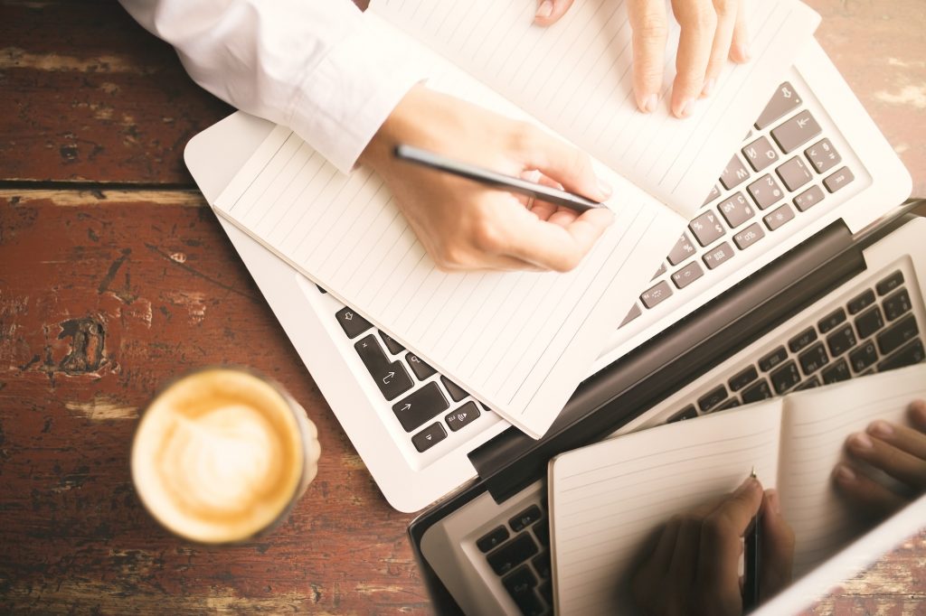 Man hands writing in the diary, coffee mug and laptop on wooden