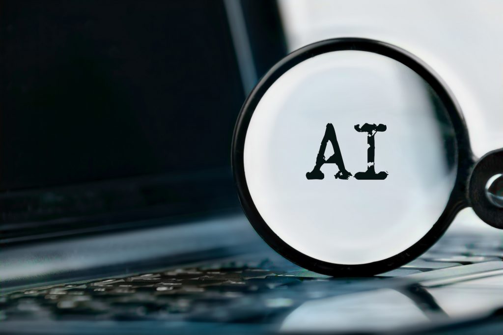  A magnifying glass over a laptop keyboard with the word AI in the center of the magnifying glass.